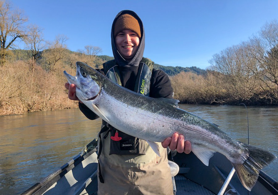 A person wearing a brown beanie, black hoodie, and waders smiles while holding a large fish in both hands. They are standing on a boat near Tillamook with leafless trees and a blue sky in the background. The water's surface is calm, reflecting the natural surroundings of Oregon.