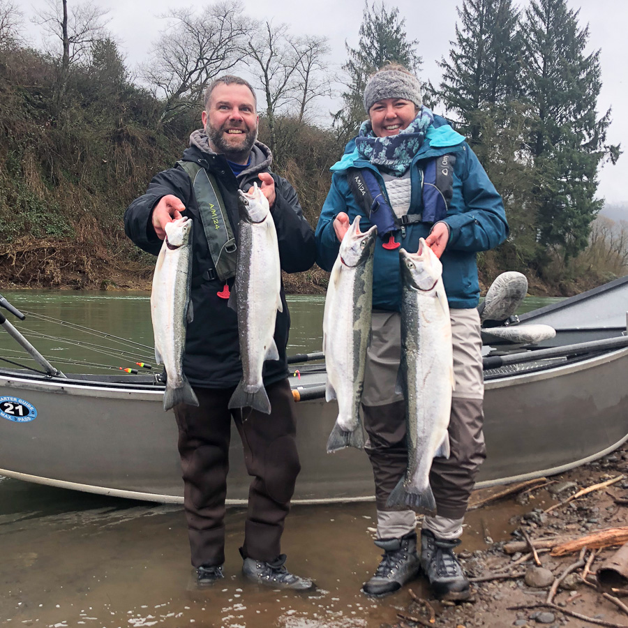 A man and a woman stand on the banks of the Wilson River in front of a Drift boat holding four winter steelhead they caught on a guided fishing trip