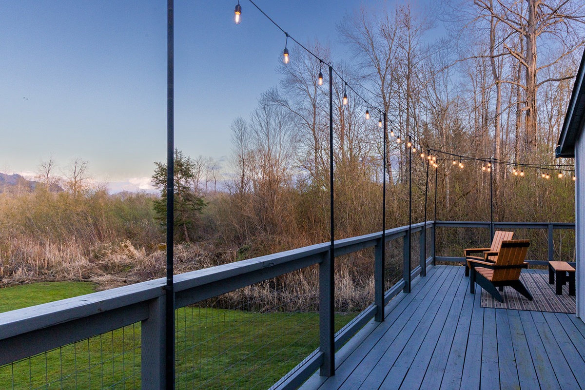 A spacious wooden deck with string lights hanging above features two wooden Adirondack chairs. The deck overlooks a serene landscape with a grassy yard and bare trees, suggesting late autumn or early spring. The sky is clear with a subtle gradient from blue to light at the horizon, perfect for an Airbnb vacation rental in Cathlamet.