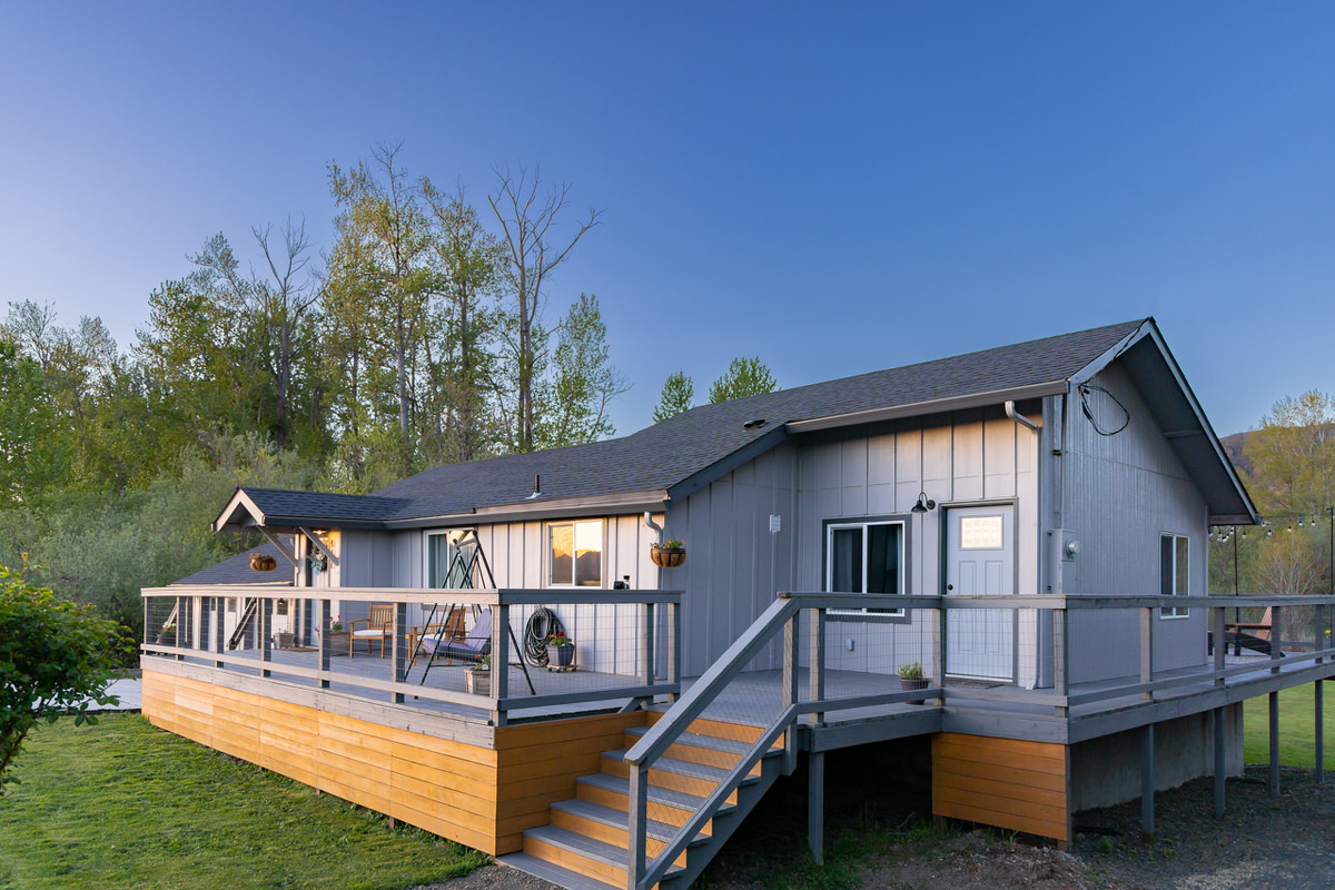 A modern single-story gray house with a gable roof on Puget Island is depicted. It features a large wooden deck with railings, stairs, and potted plants. The Airbnb has large windows and a door with a small awning. It's set against a backdrop of green trees under a clear, blue sky in the evening light.