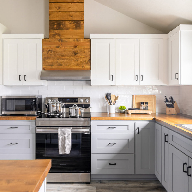 Modern kitchen with white upper cabinets and light blue lower cabinets. A stainless steel stove sits in the center below a wooden vent hood. The countertop is wooden with utensils, a cutting board, and a knife block. A microwave is on the left, with a white subway tile backsplash throughout.