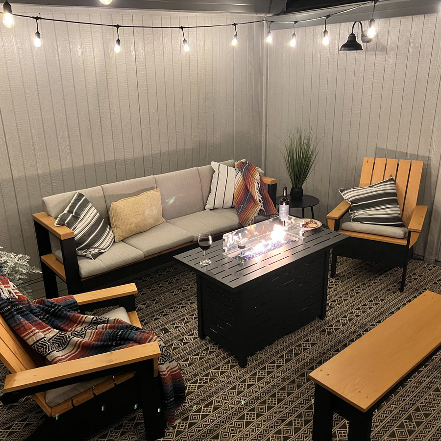 A cozy outdoor seating area features a gray cushioned sectional sofa, two wooden chairs with striped cushions, and a black rectangular fire pit table. String lights hang overhead, and a patterned rug covers the floor. A multicolored blanket, wine glasses, and a potted plant add to the ambiance.