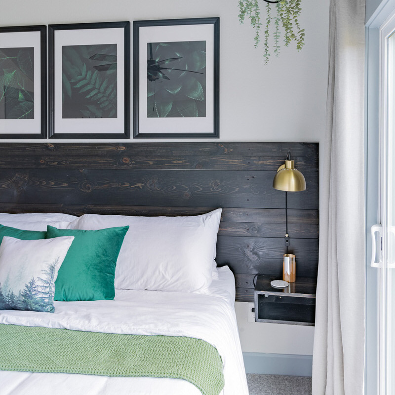 A modern bedroom features a bed with white linens and green accent pillows. Above the bed, there's a dark wooden headboard, and three framed botanical prints. A gold lamp and a small shelf holding a few items are mounted on the headboard. A window with gray curtains is on the right.